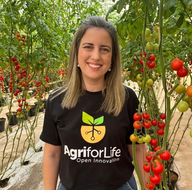 Foto de Maira, com cabelos longos loiros, vestindo camisa preta em uma estufa, rodeada por tomates, simbolizando o cultivo e a agricultura sustentável.